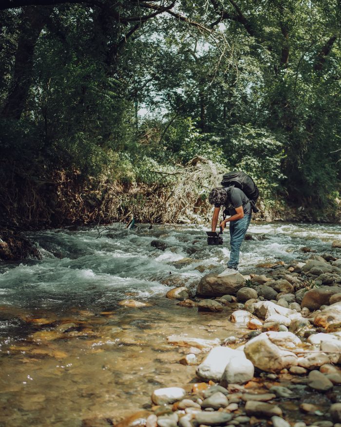 about-us A videographer films a scenic river amidst lush green trees, showcasing nature's beauty.
