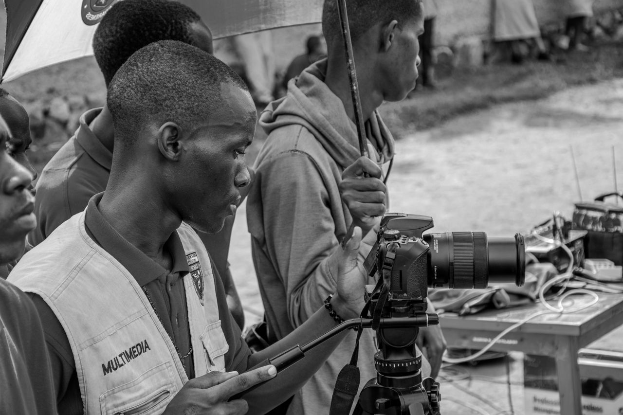 Black and white image of a filmmaker setting up a camera outdoors, capturing a documentary scene.